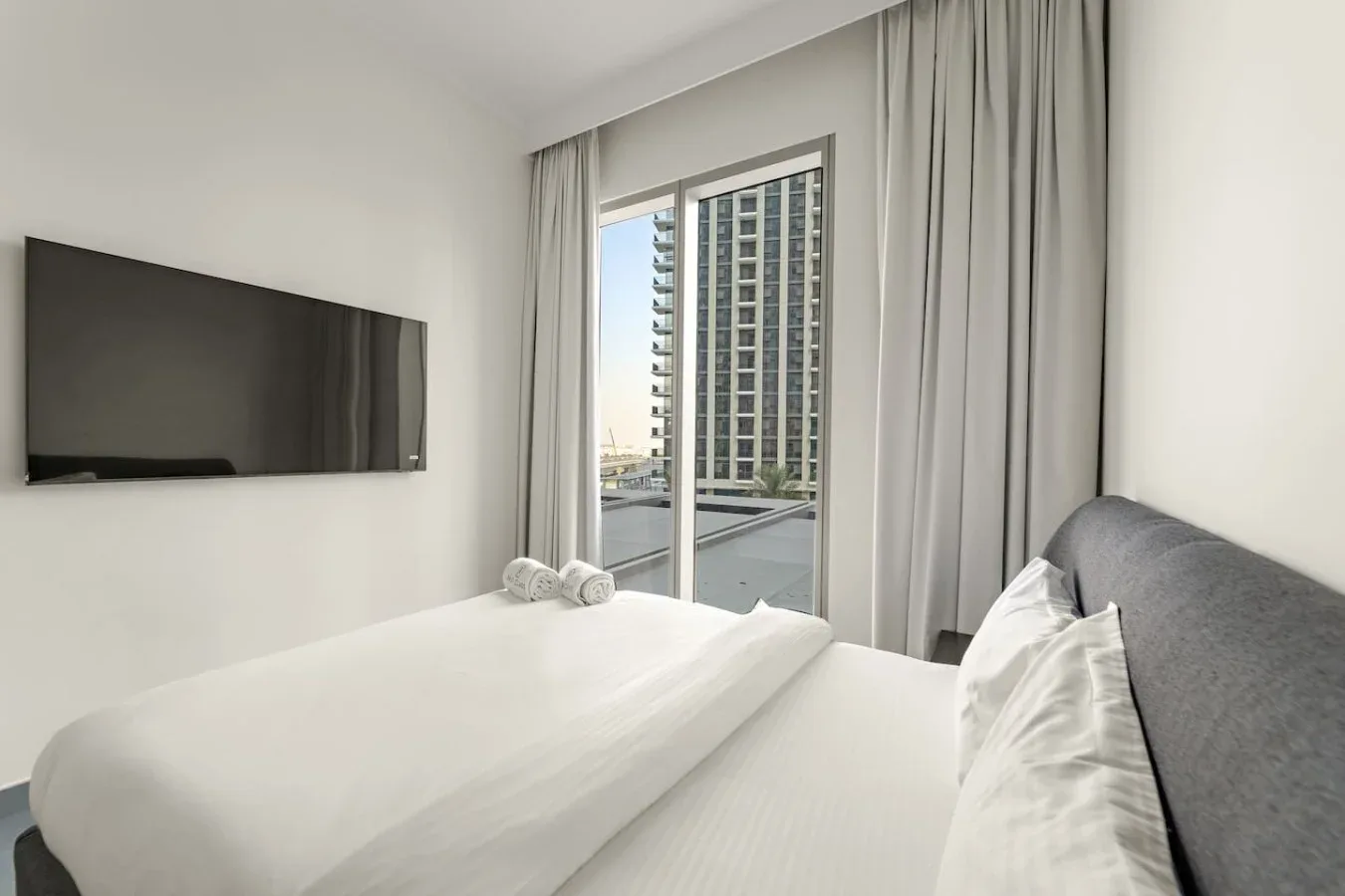 A bedroom with a bed beside a large window overlooking nearby buildings.