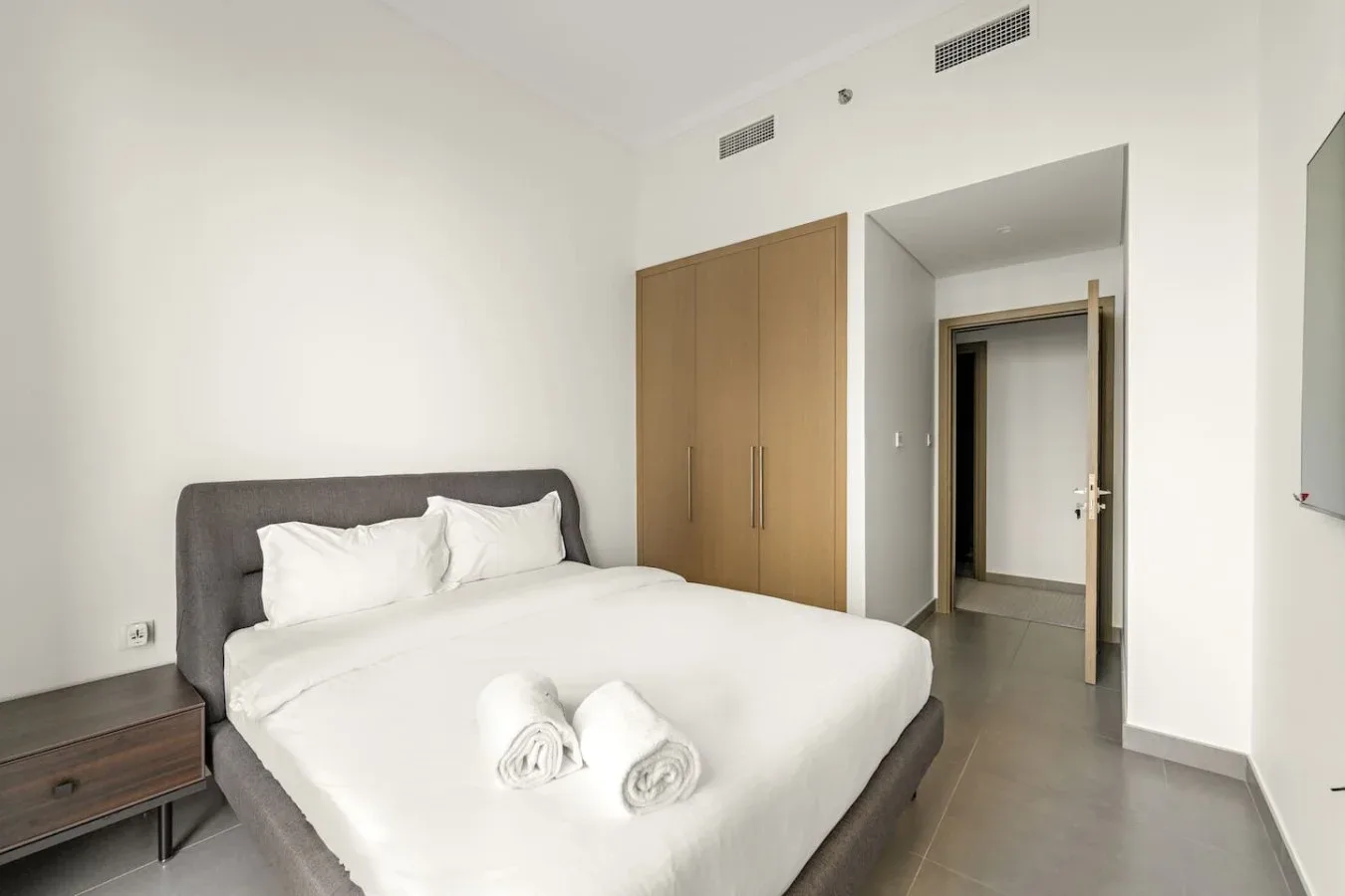 A bedroom with a neatly made bed and towels, beside a wooden wardrobe.