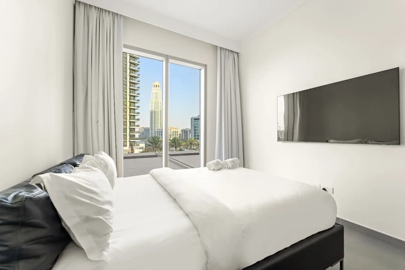 A bright bedroom with white bedding, a wall-mounted television, and a city view.