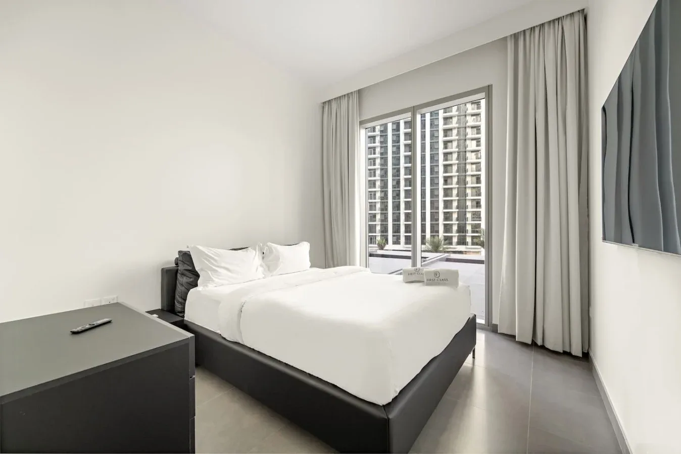A bright bedroom with a neatly made bed and a large curtained window.
