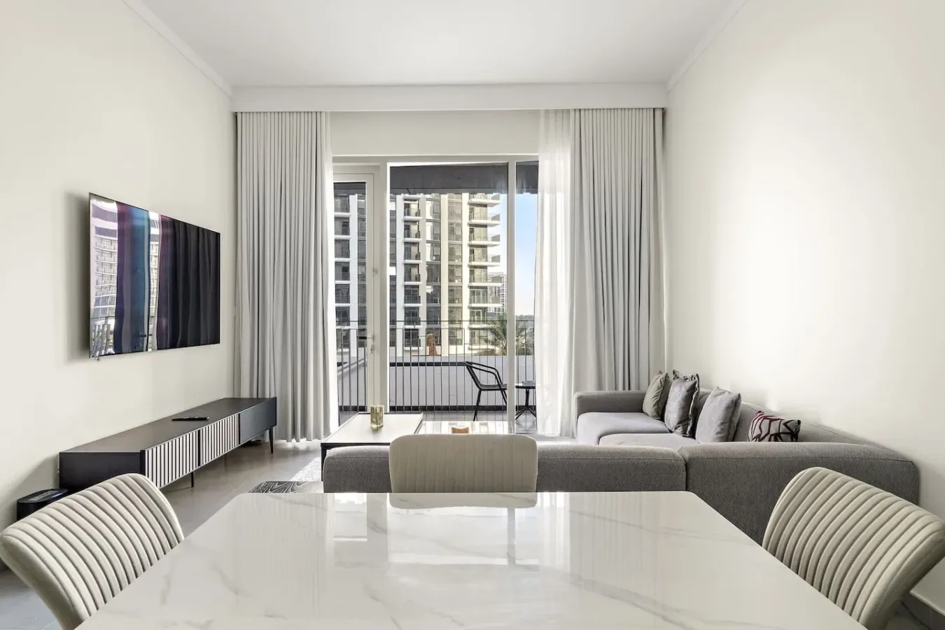 A bright living room with gray seating, a wall-mounted television, and large windows.