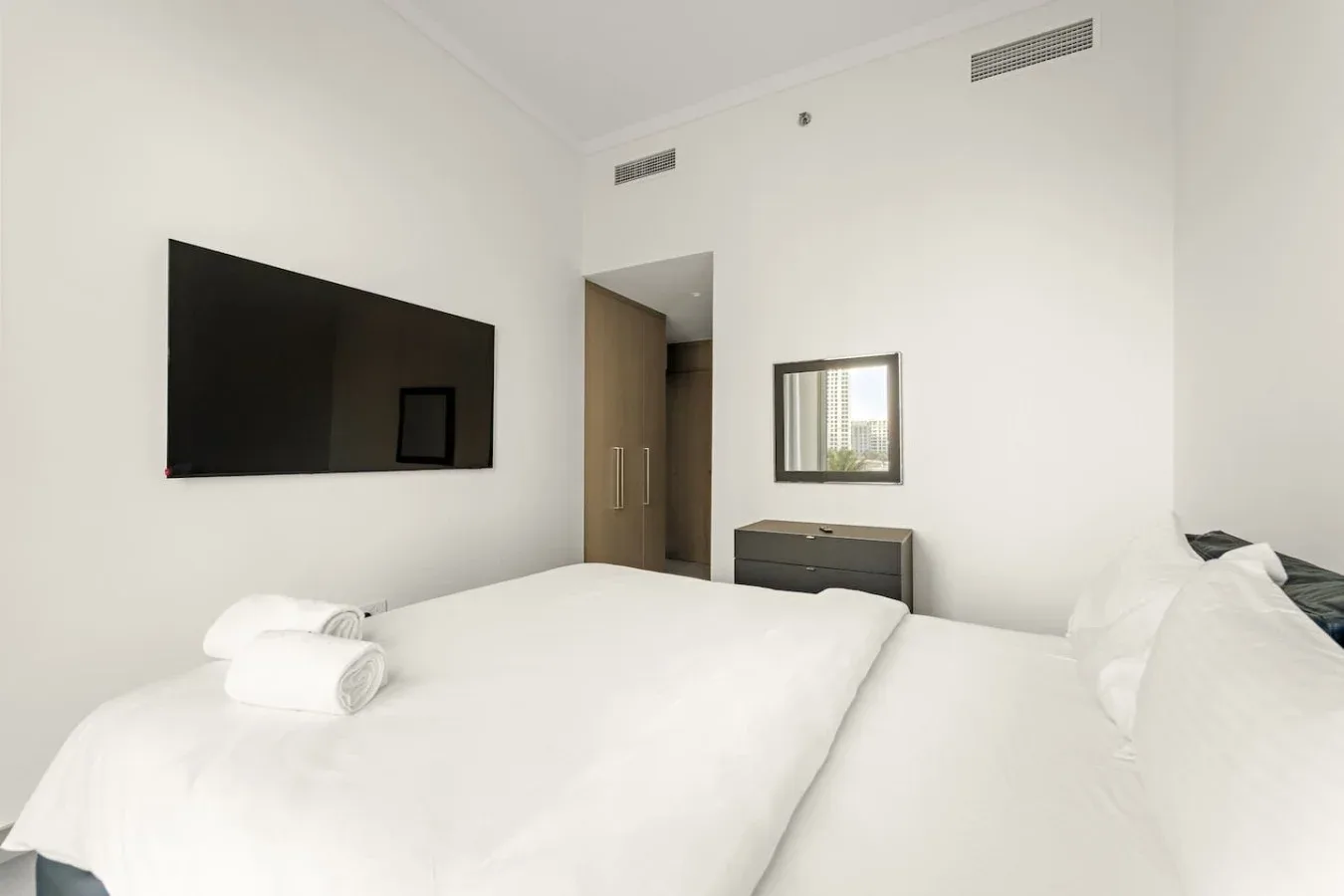 A bright bedroom with white bedding, folded towels, and a wall-mounted television.