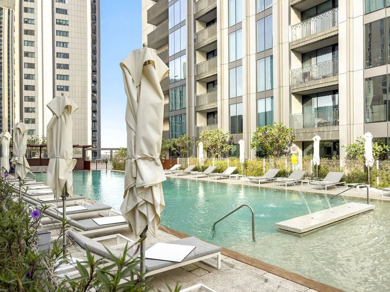 An outdoor swimming pool with lounge seating and closed umbrellas beside modern buildings.