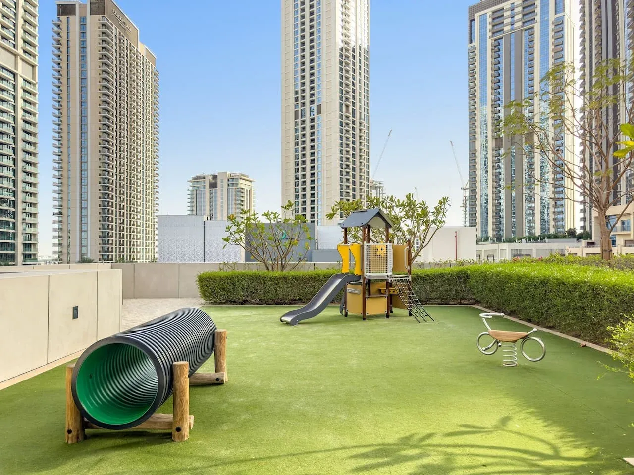 An outdoor playground with a slide, tunnel, and green play surface among high-rise buildings.