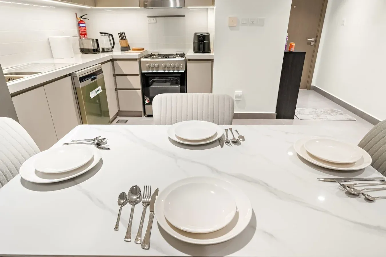 A dining table set with plates and cutlery stands in front of a compact kitchen.