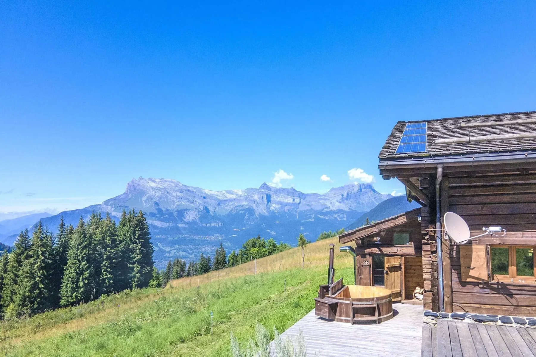A wooden chalet and deck overlook a grassy hillside, trees, and distant mountains.