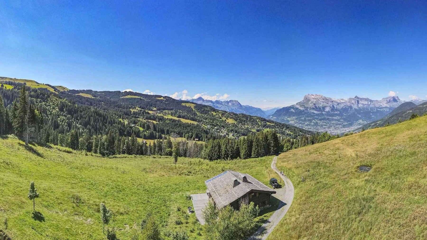A chalet sits in a grassy alpine landscape with forested hills and distant mountains.