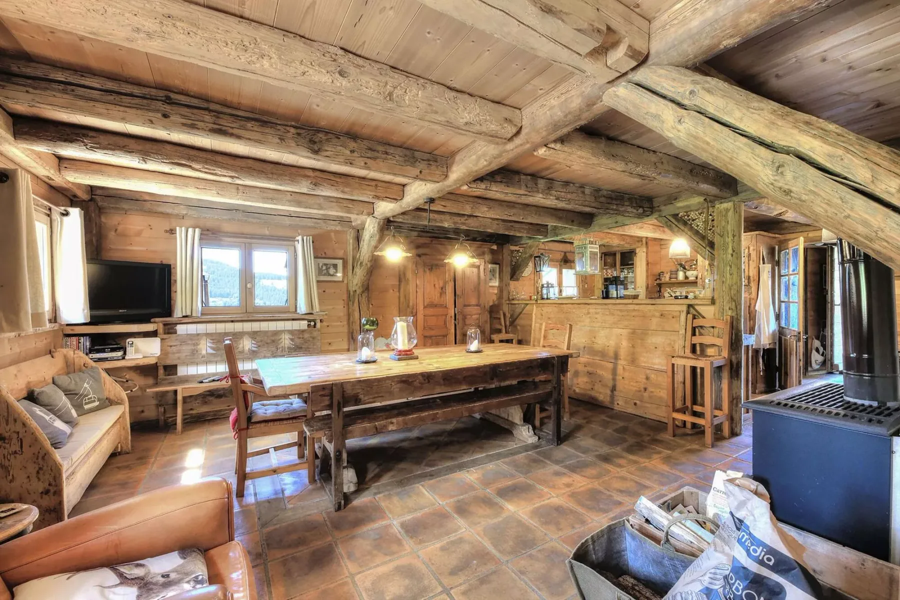 A rustic dining and living area with a long wooden table beneath exposed timber beams.