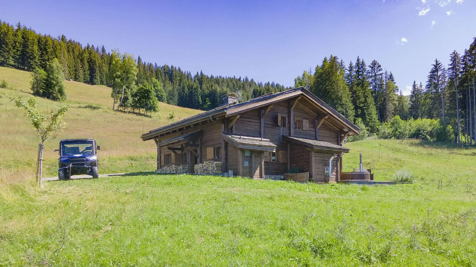 A wooden chalet stands in a grassy meadow with forested hills behind it.
