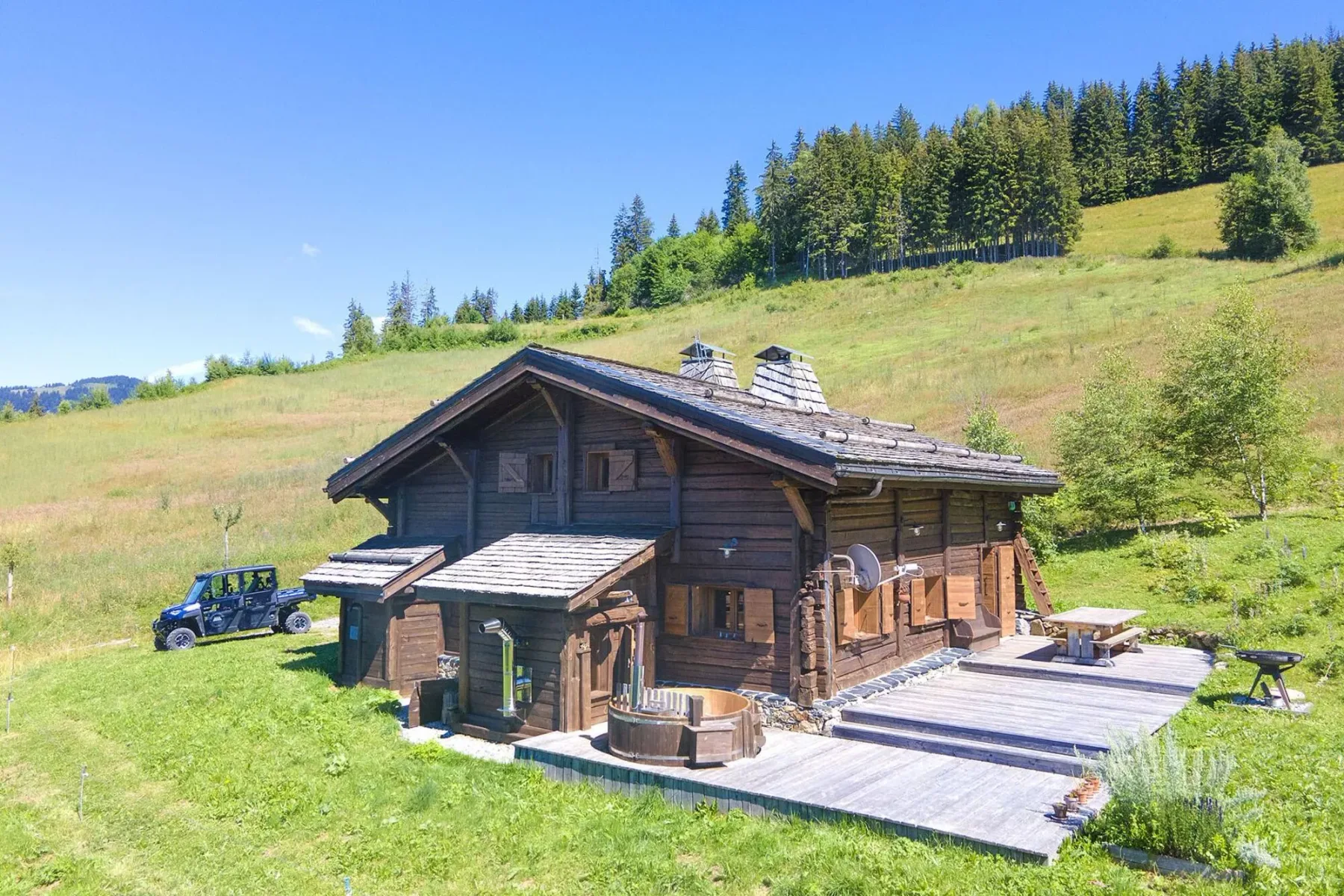 A wooden chalet sits in a grassy hillside clearing beside a broad deck.