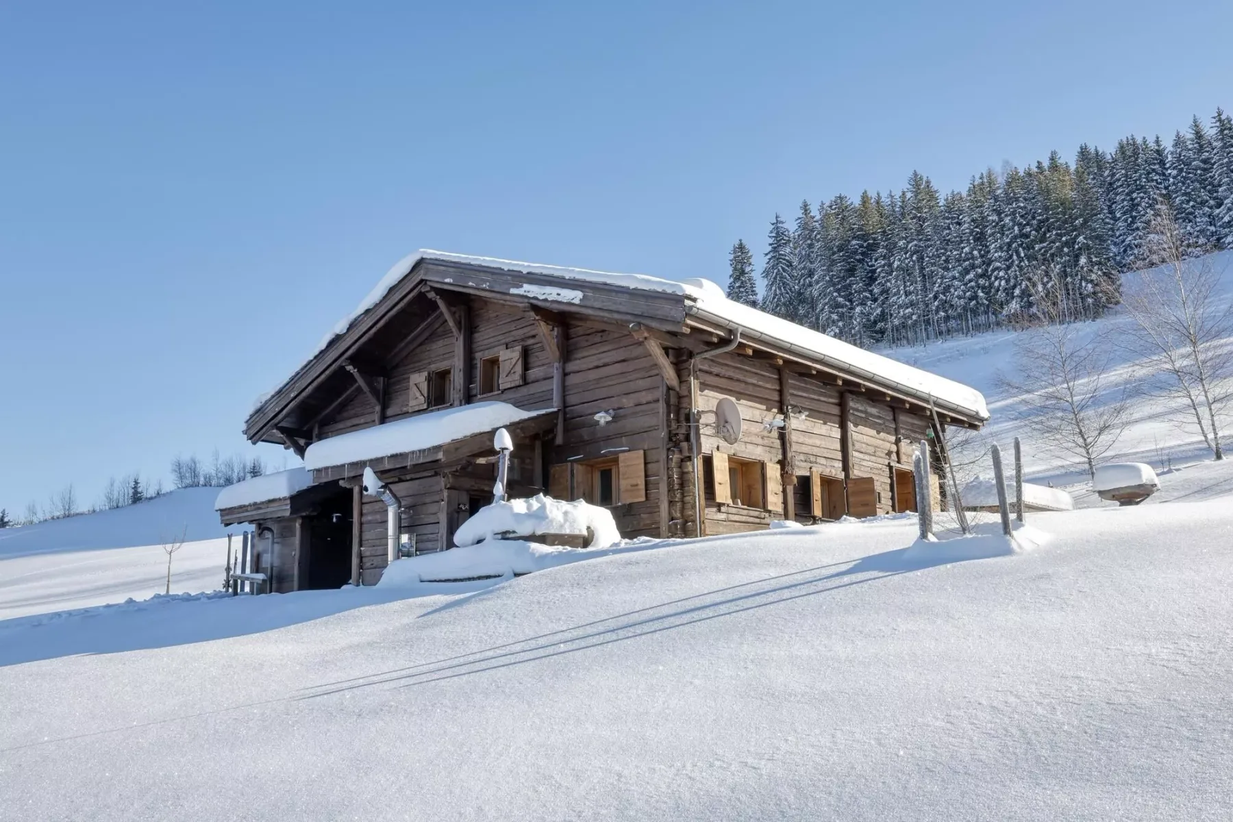 A wooden chalet stands beside a snowy hillside under a clear blue sky.