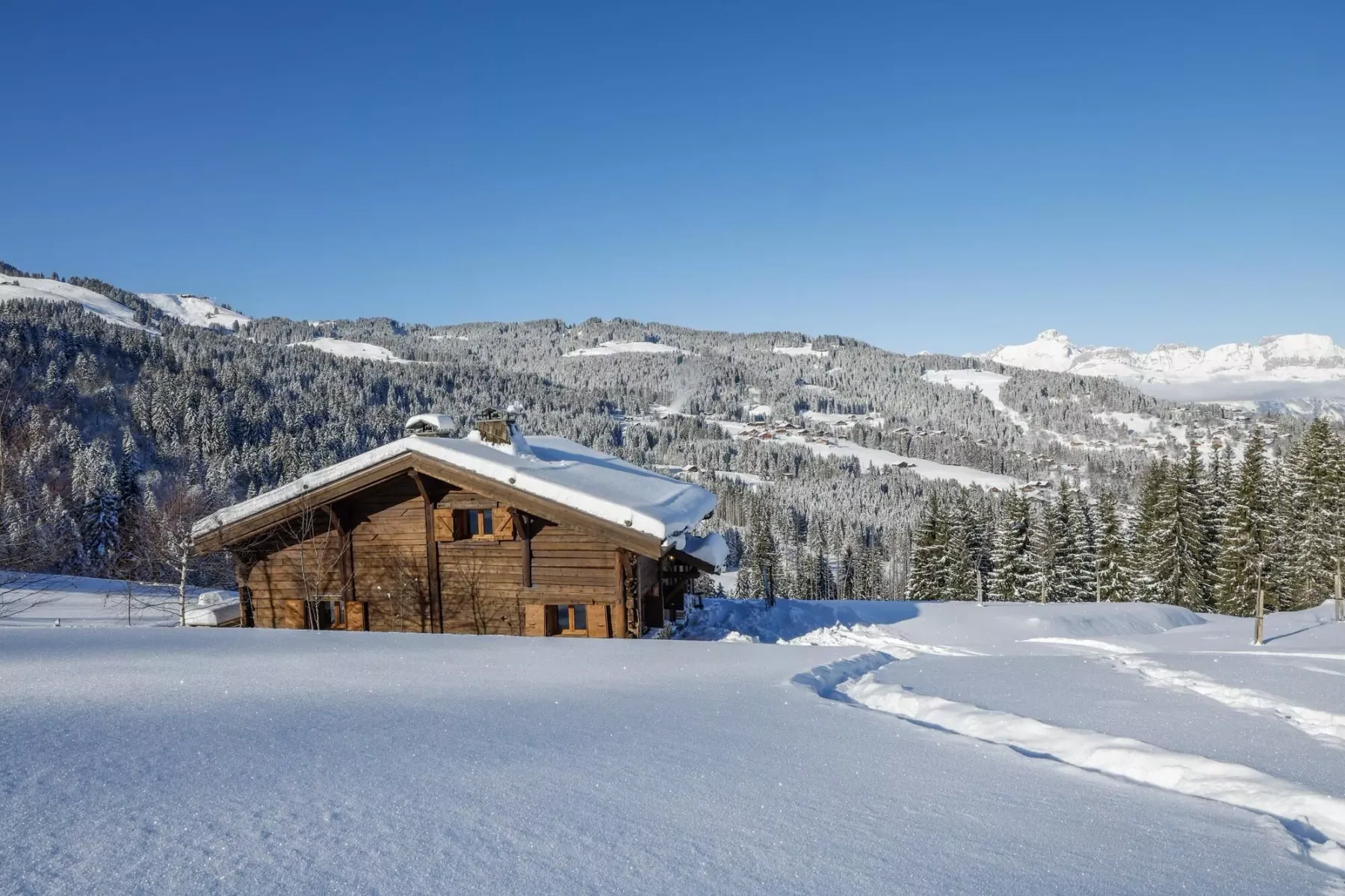 A wooden chalet sits in a snowy mountain landscape beneath a clear blue sky.