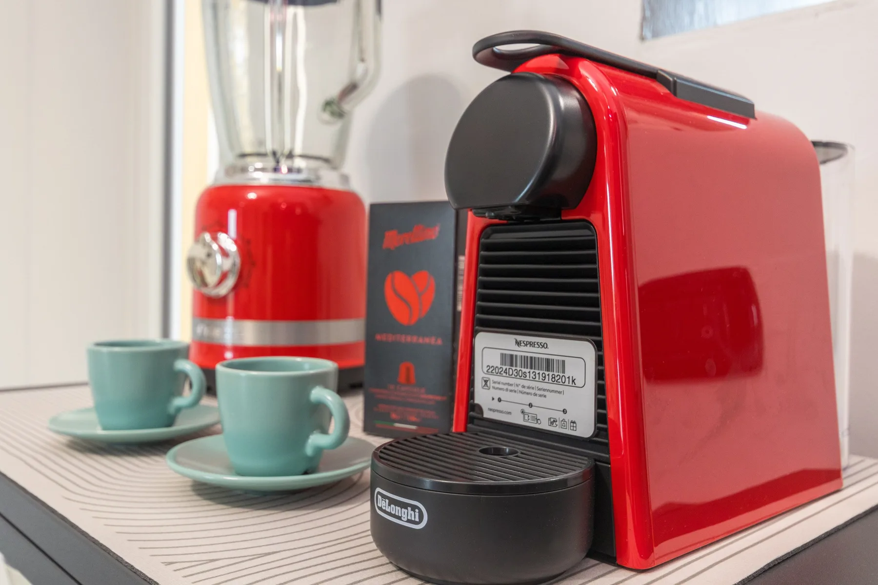 Una cafetera roja y una licuadora están junto a dos tazas sobre una encimera.