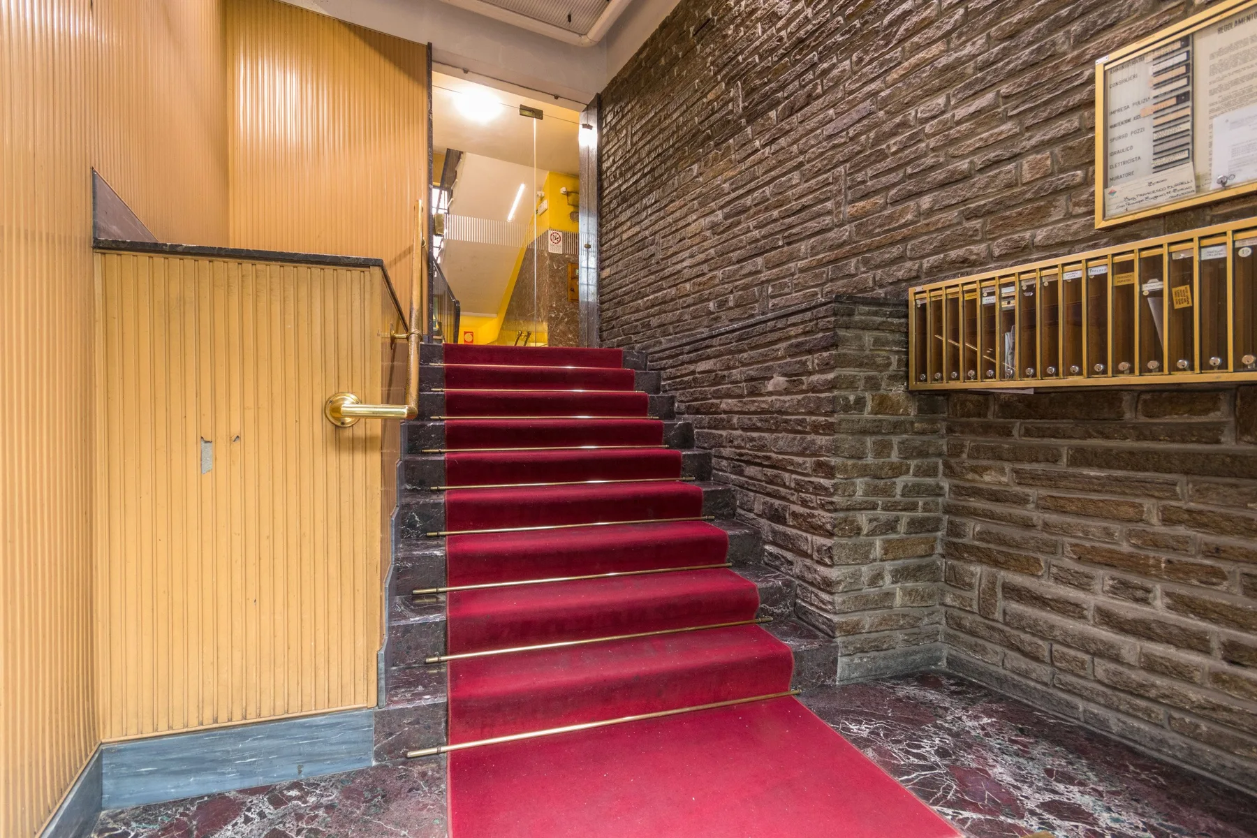Una escalera alfombrada de rojo sube entre paneles de madera y una pared de ladrillo.