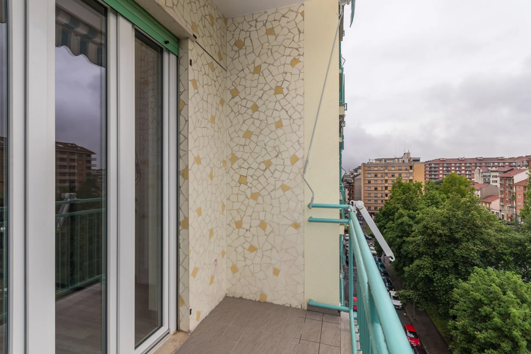 Un balcón estrecho con puertas corredizas de vidrio da a árboles y edificios de apartamentos cercanos.