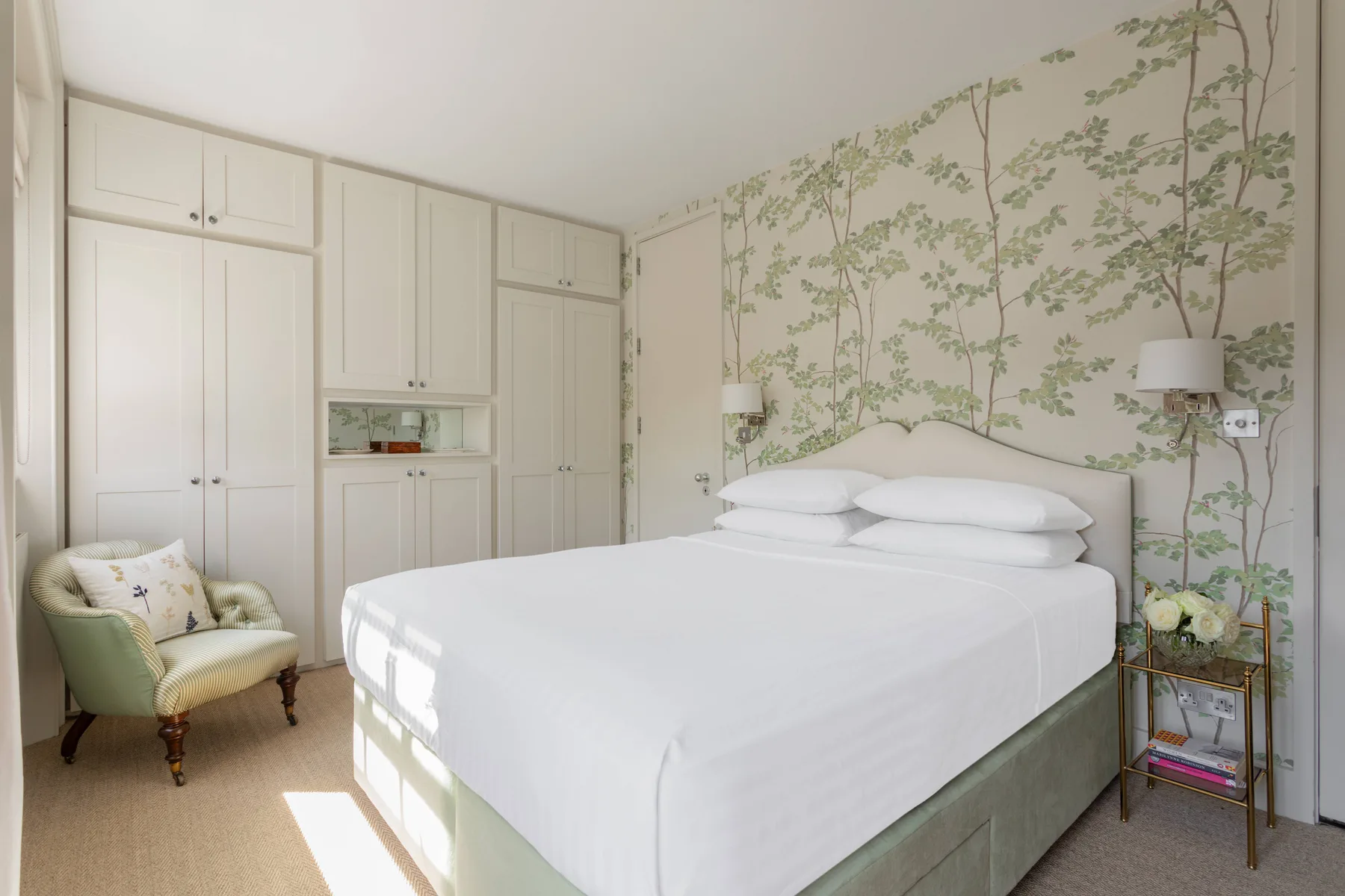 A bright bedroom with a large bed, built-in wardrobes, and leafy wallpaper.