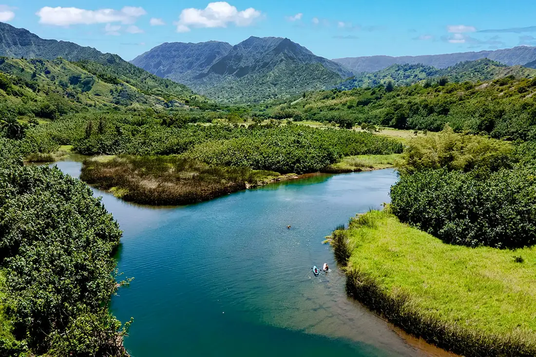 A river winds through green wetlands beneath a range of forested mountains.