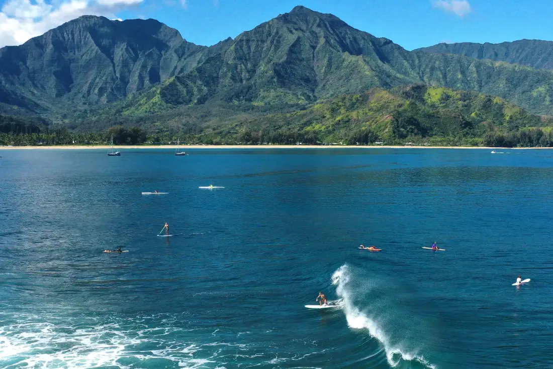 A mountain coastline rises behind blue ocean water, with surfers riding small waves.