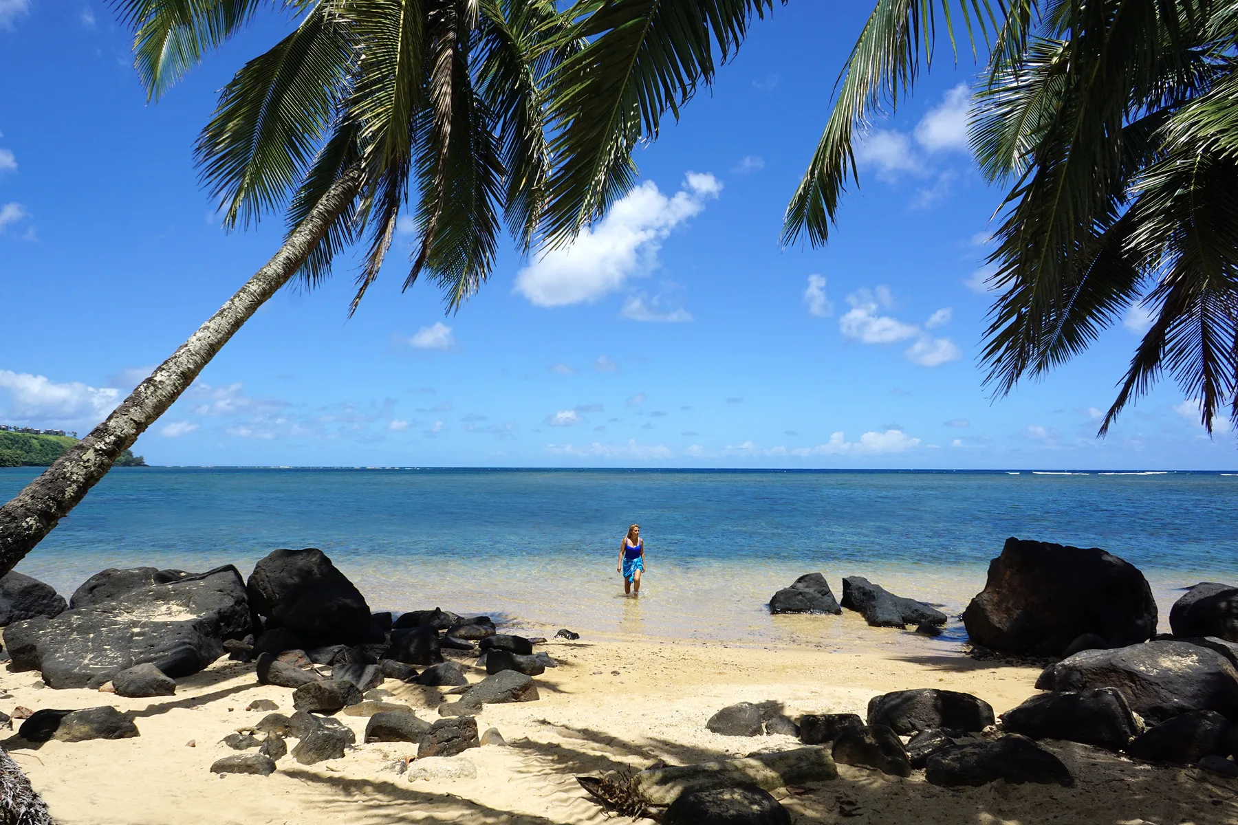 A sandy beach with dark rocks, palm fronds, and a person standing by the water.