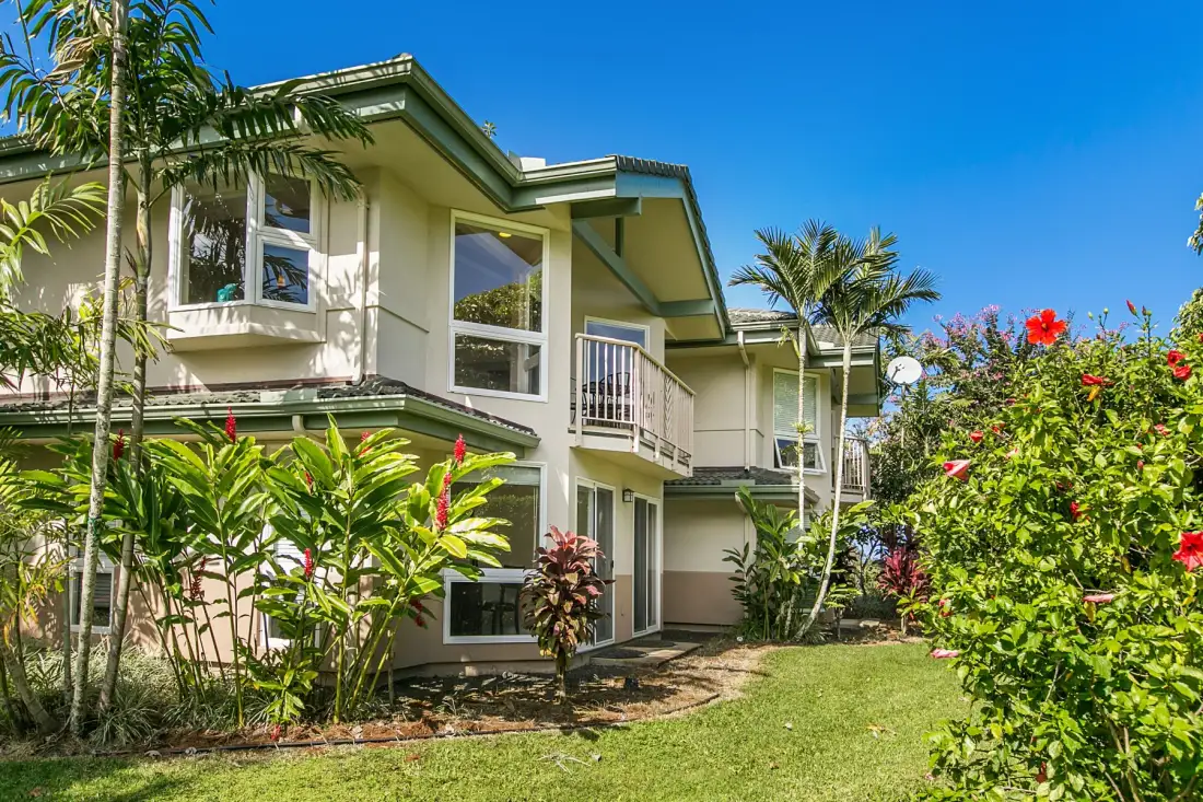 A two-story residential building stands amid tropical plants and a green lawn.