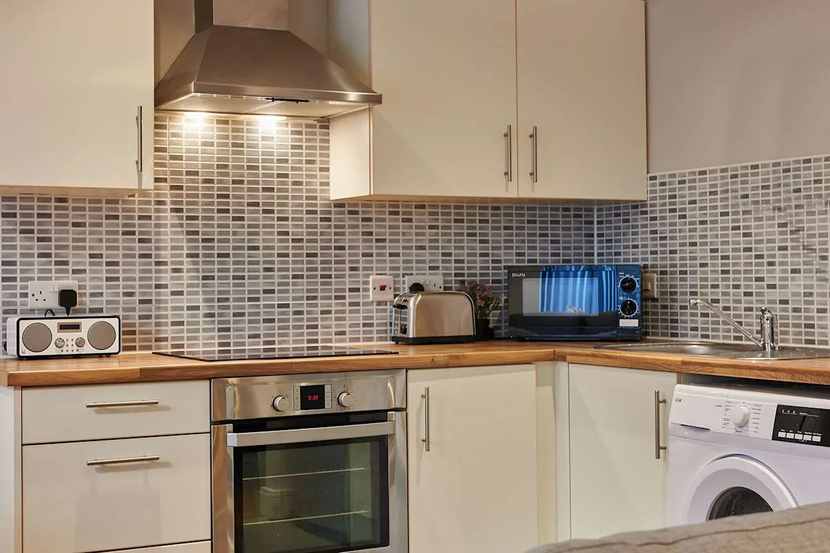 Una cocina compacta con gabinetes color crema, salpicadero de azulejos y electrodomésticos sobre la encimera.