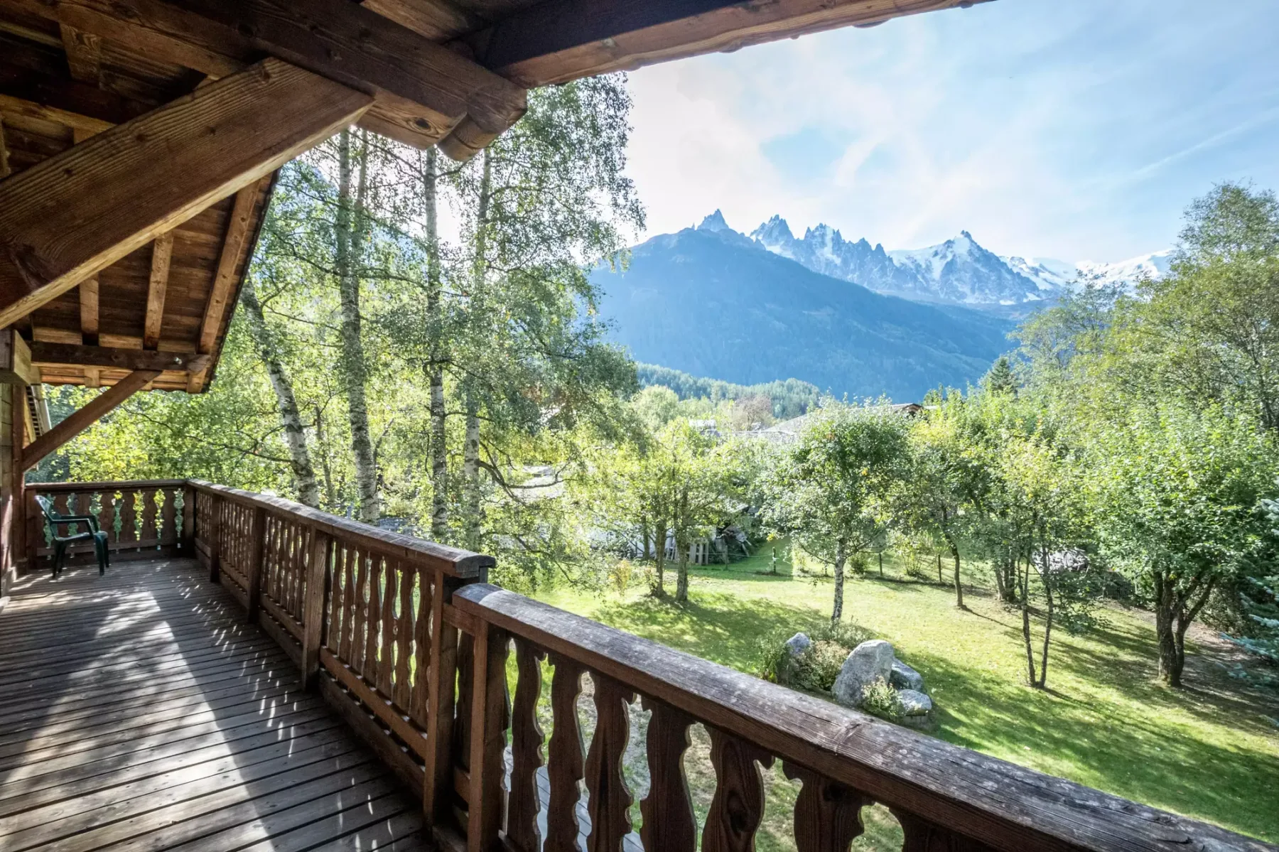 A covered wooden balcony overlooks a green orchard and distant mountain peaks.