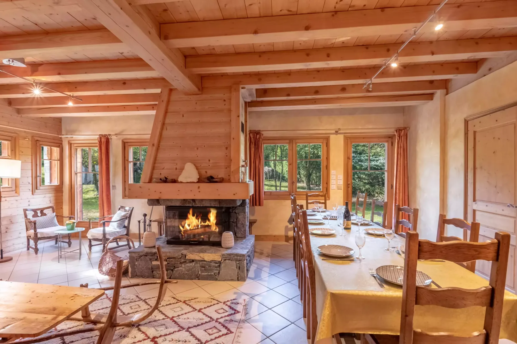 A wood-beamed dining and living room with a long dining table and fireplace.