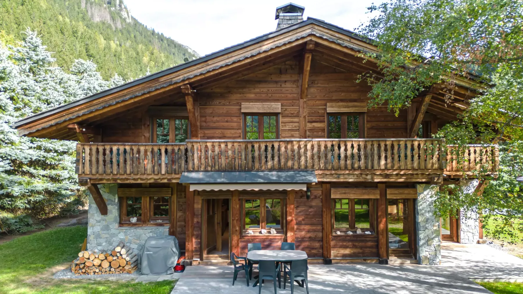 A wooden chalet with a balcony and outdoor seating in a green mountain setting.