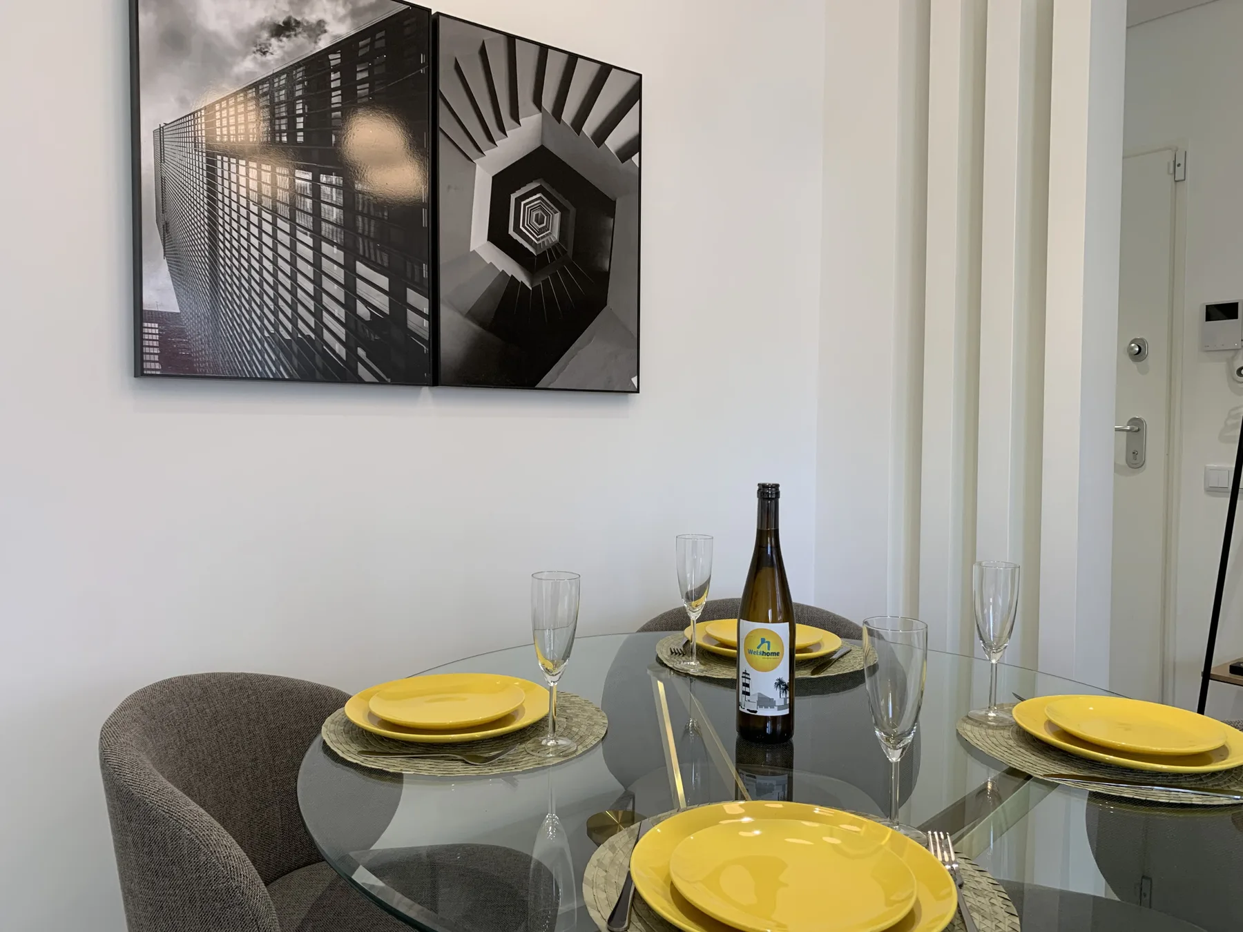 Comedor con una mesa de vidrio preparada con platos y copas bajo un cuadro en la pared.