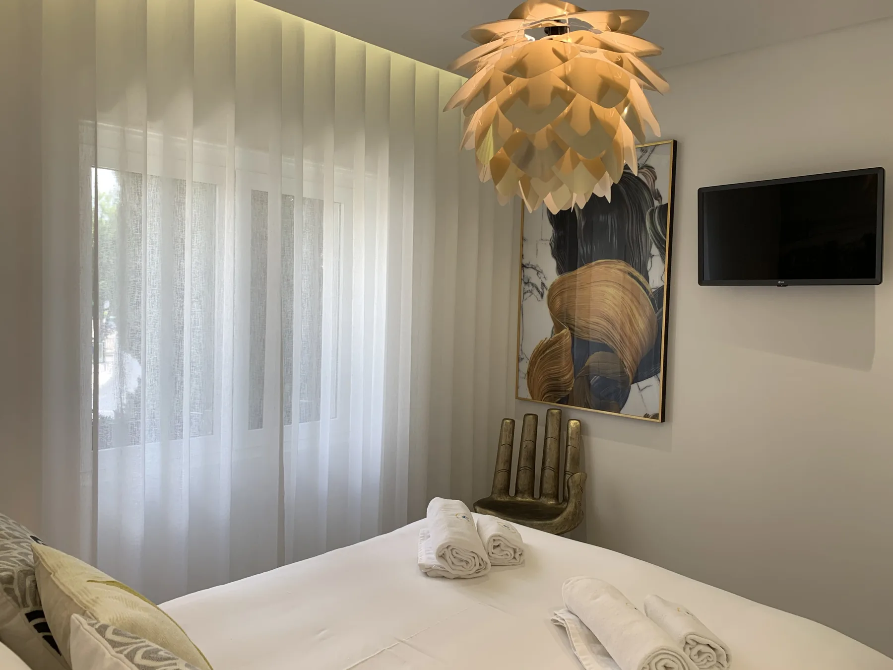 Un dormitorio con la cama bien hecha, cortinas translúcidas y un televisor en la pared.