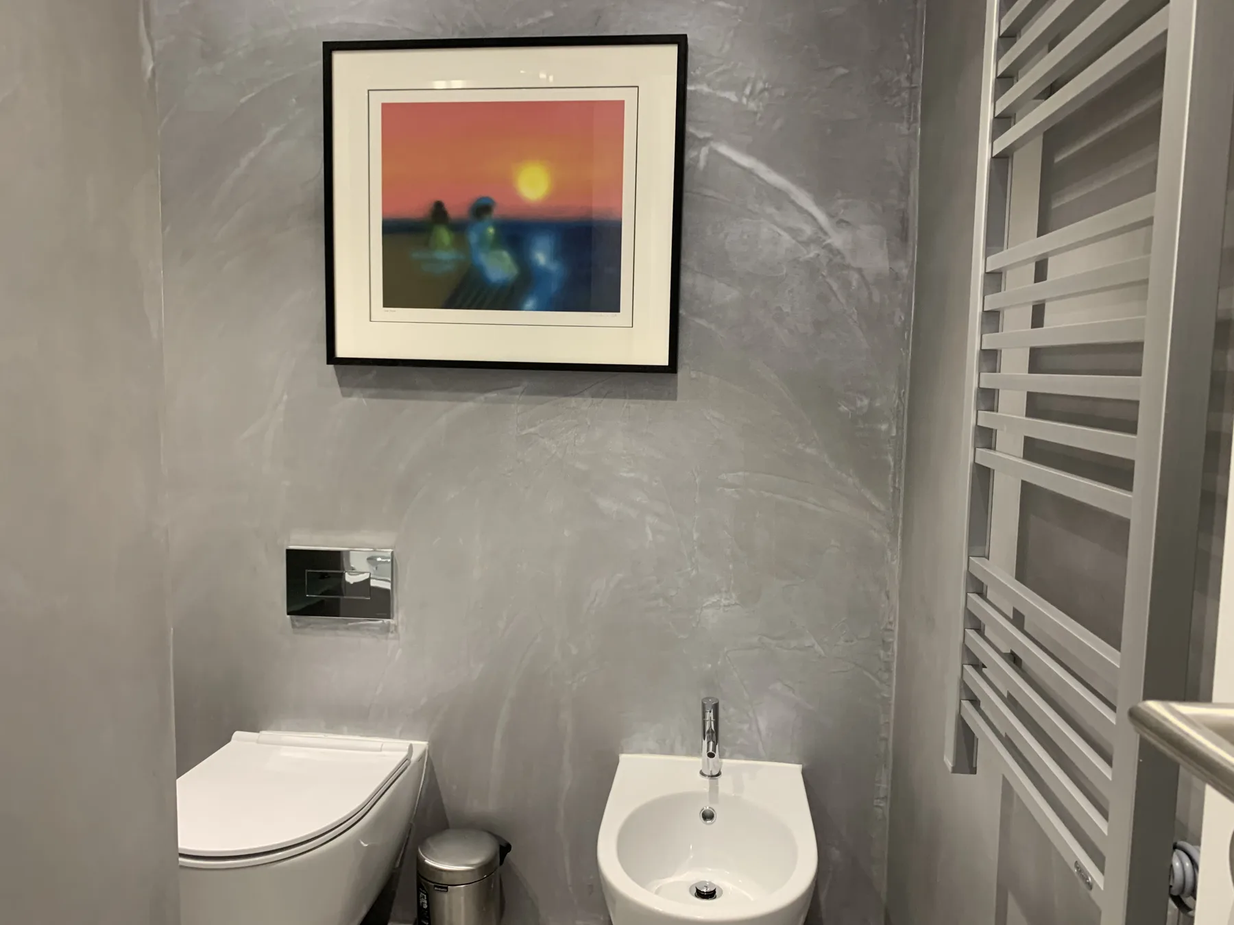 Un baño gris con un inodoro suspendido y un bidé bajo una obra enmarcada.