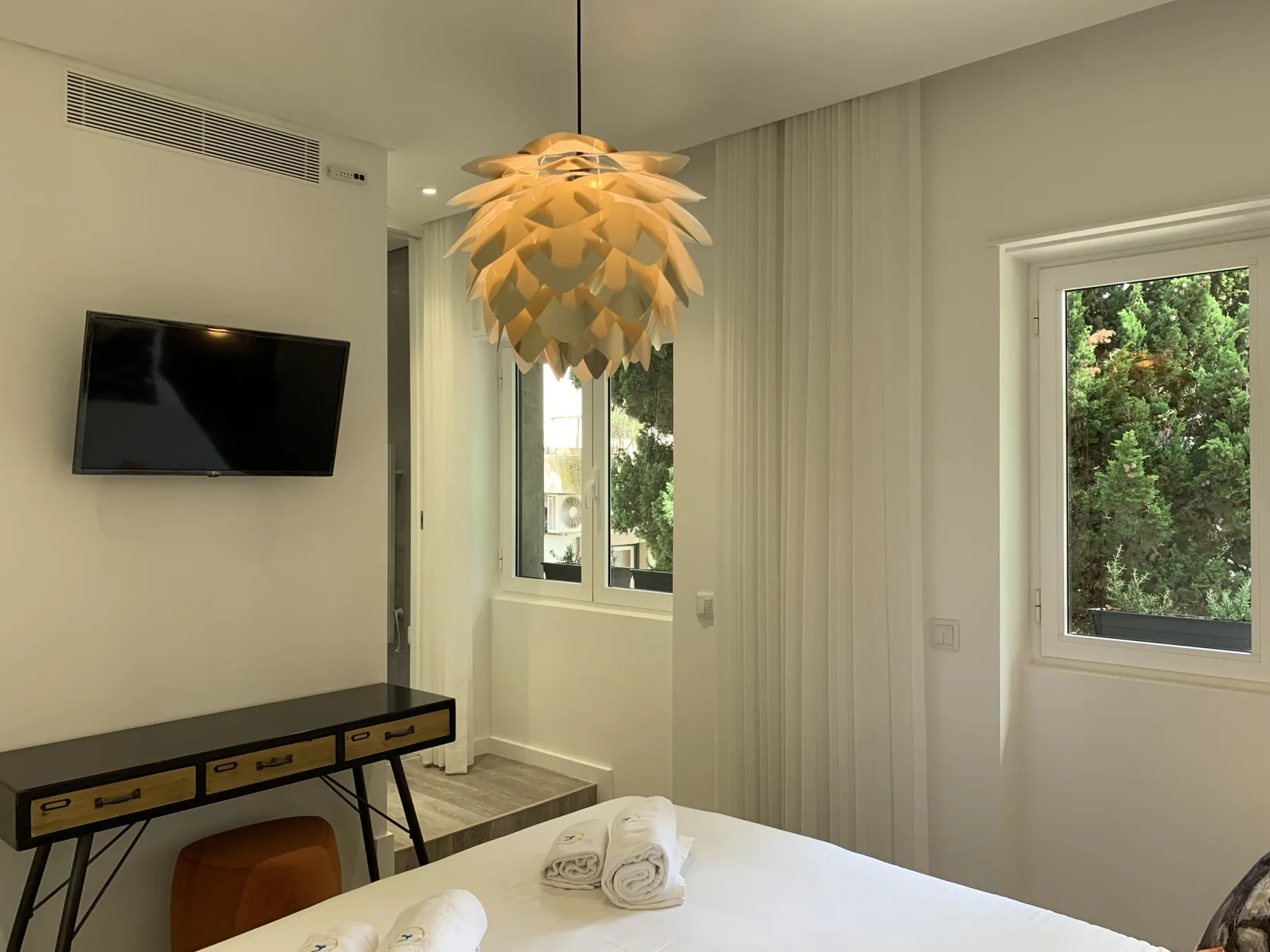 Dormitorio luminoso con una cama, un televisor en la pared y ventanas con vistas a la vegetación.