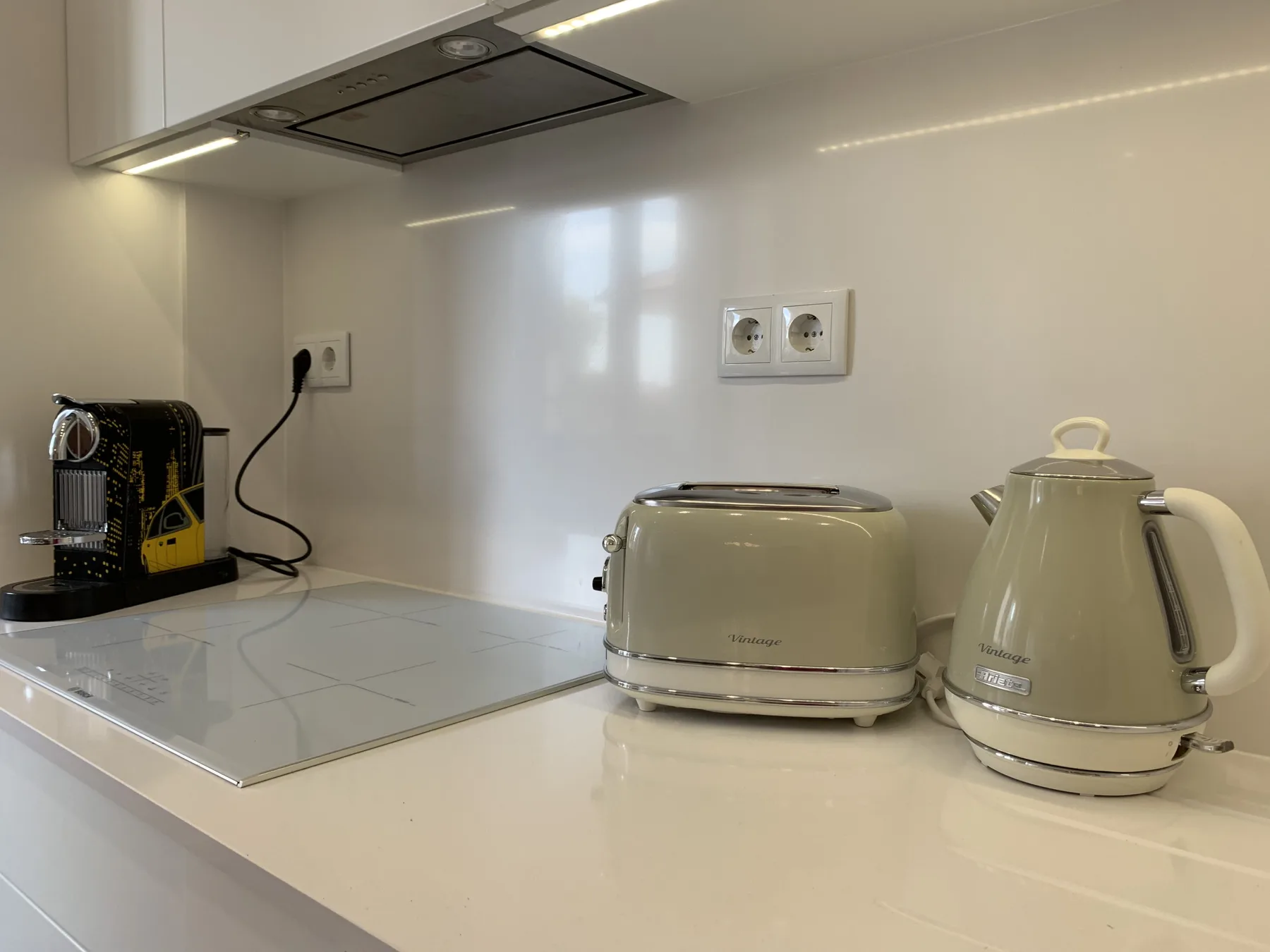 Una encimera de cocina tiene una cafetera, una tostadora y un hervidor eléctrico bajo una campana extractora.