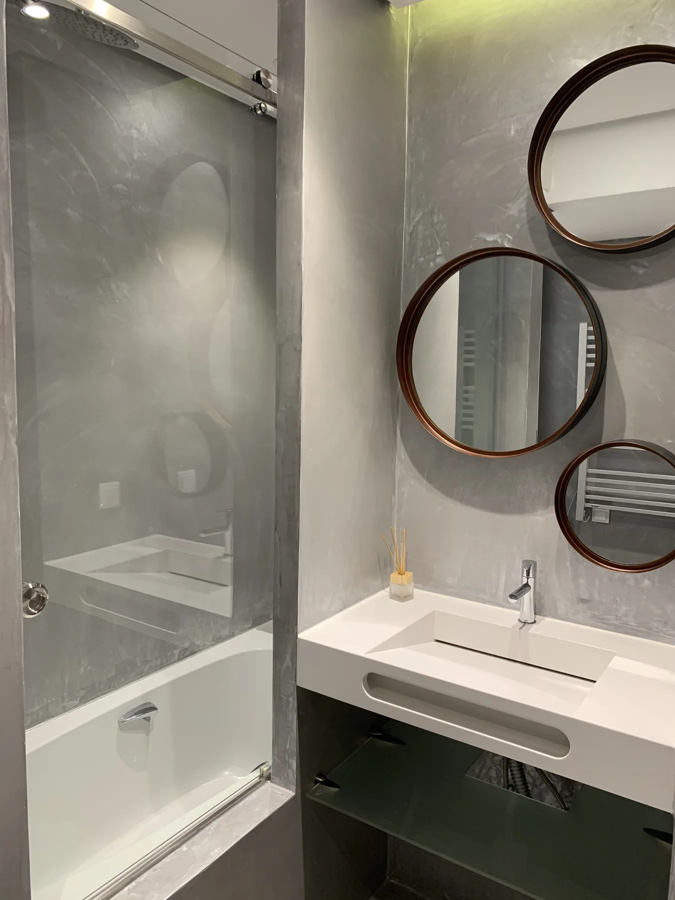 Un baño con ducha de vidrio y lavabo blanco bajo espejos redondos.