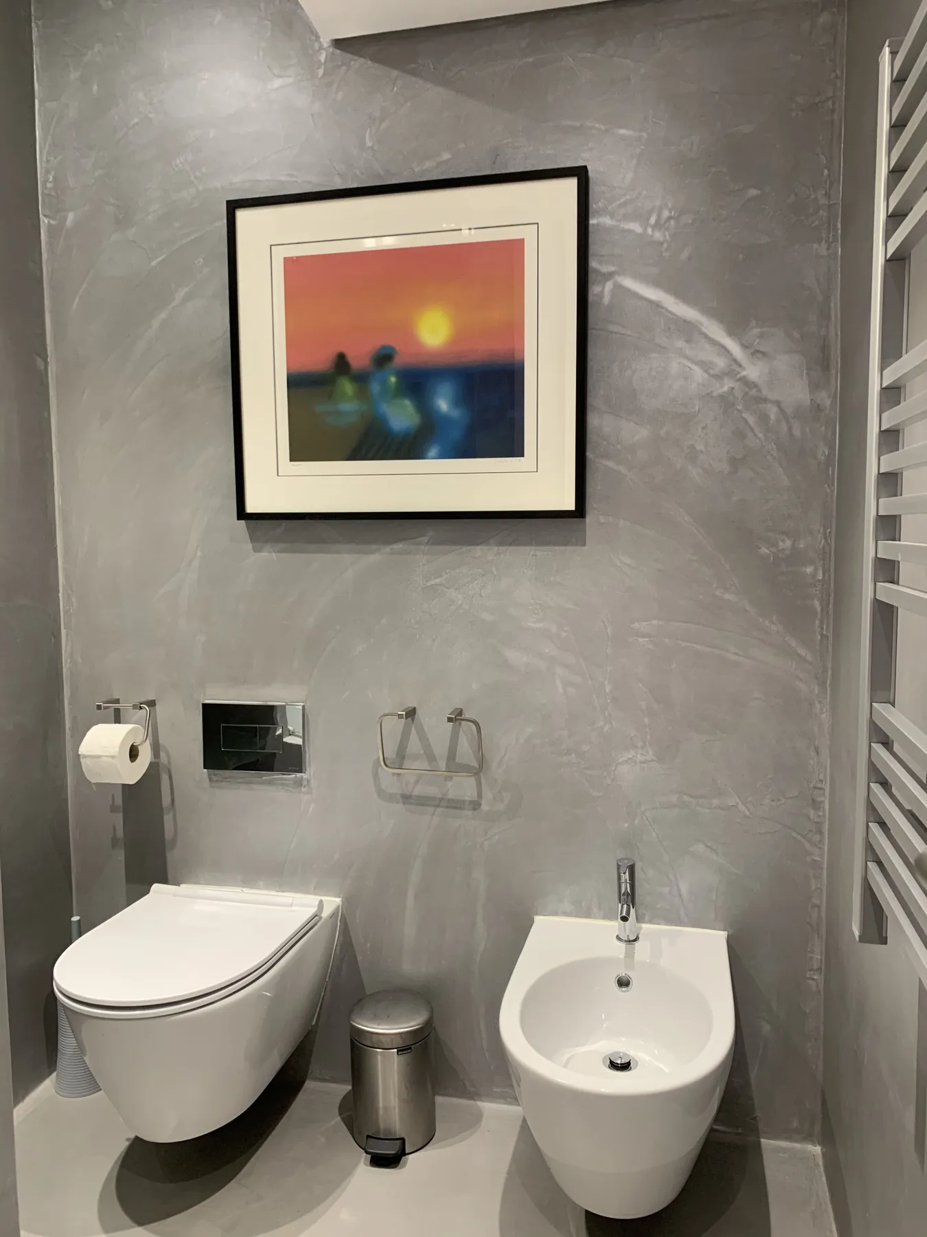 Un baño con un inodoro suspendido y un bidé bajo una obra enmarcada.