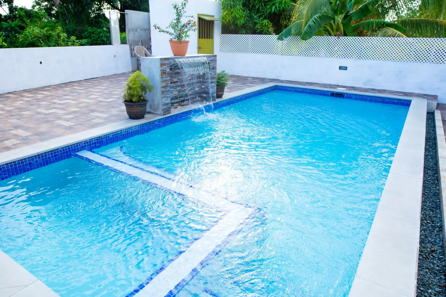 Piscina al aire libre junto a un patio con baldosas y vegetación.