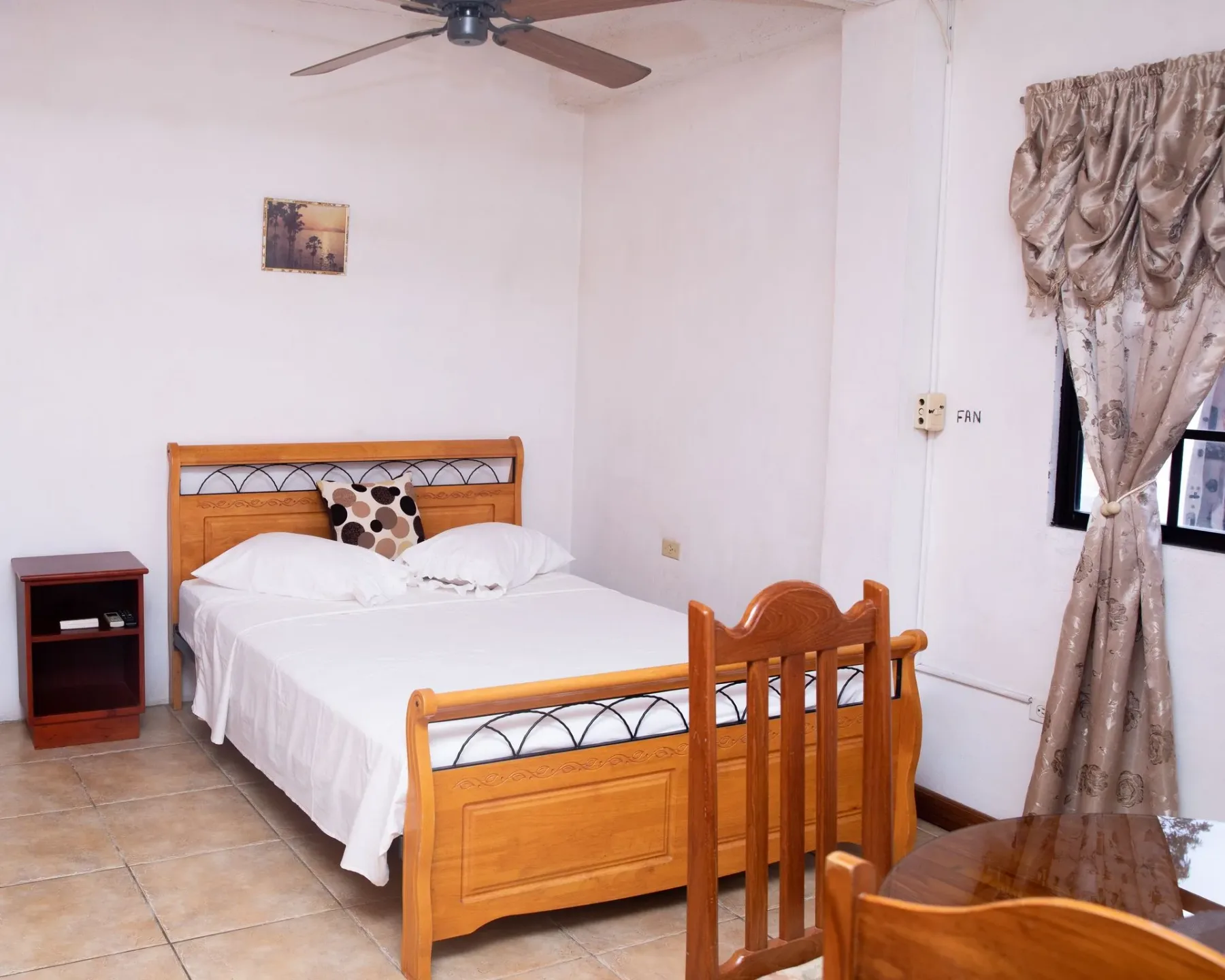 Dormitorio con una cama de madera, ventilador de techo, suelo de baldosas y una ventana con cortinas.
