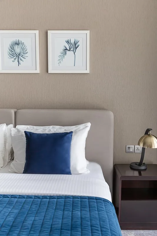 Una cama bien tendida, con ropa de cama blanca y un cojín azul, está junto a una pared color topo.