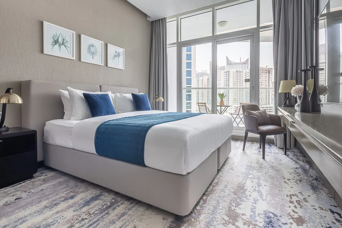 Un dormitorio luminoso con una cama bien tendida y grandes ventanales con vistas a edificios.