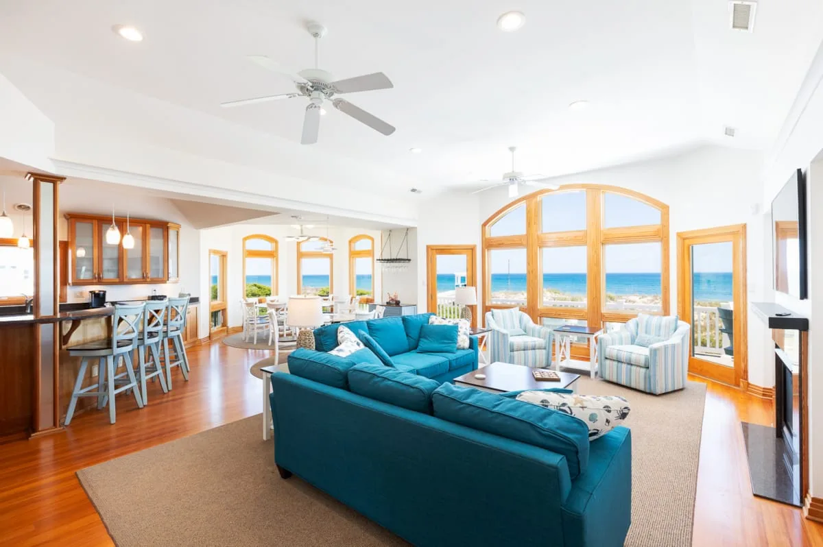 Una luminosa sala de estar con asientos color turquesa, comedor y grandes ventanales con vistas al mar.