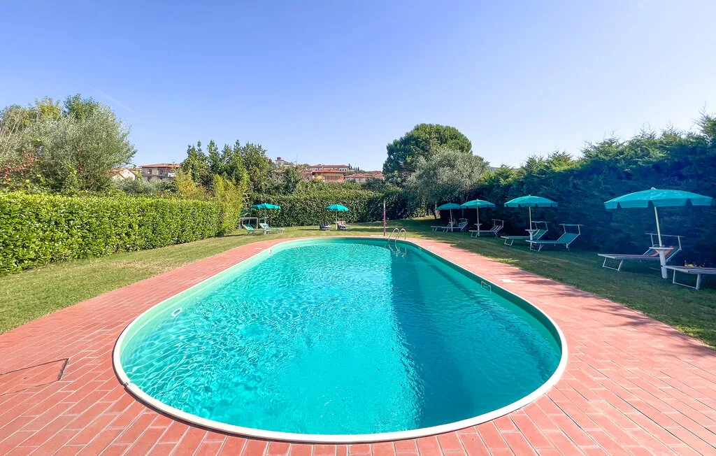 Una piscina al aire libre está rodeada por una zona pavimentada, vegetación y asientos con sombra.
