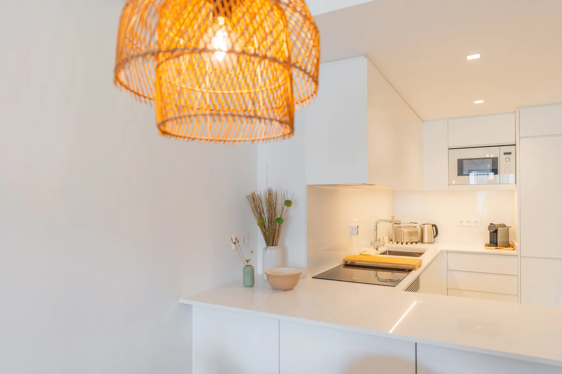 Una cocina blanca y luminosa con encimera, placa de cocina y lámpara colgante tejida.