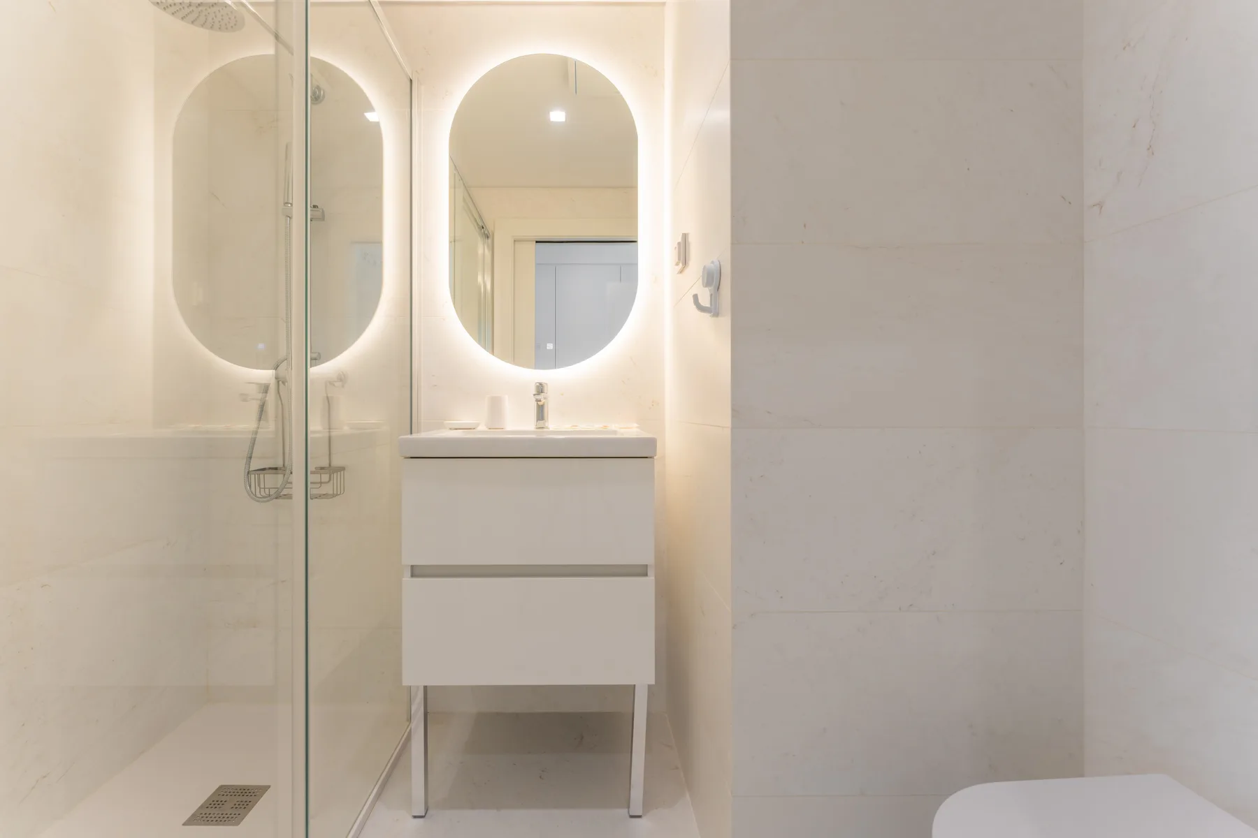 Un baño luminoso con ducha de vidrio y un mueble de lavabo bajo un espejo iluminado.