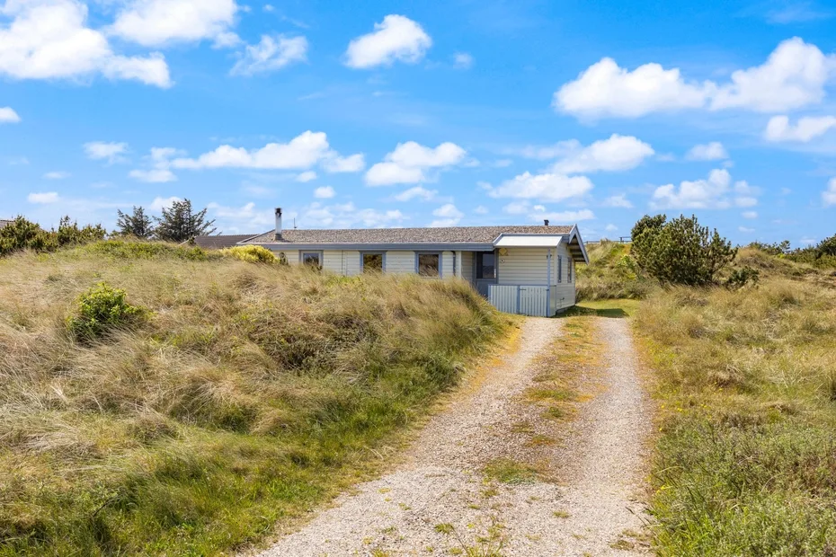 Una casa baja se encuentra al final de un camino de grava entre dunas cubiertas de hierba.