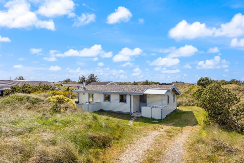 Una casa pequeña se encuentra entre dunas cubiertas de hierba bajo un cielo azul.