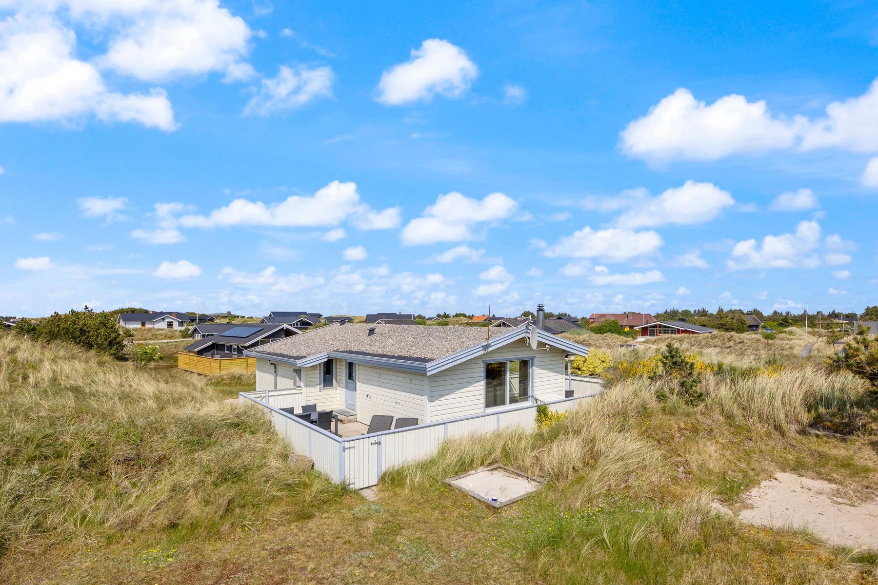 Una casa de color claro está entre dunas cubiertas de hierba bajo un cielo azul con nubes.