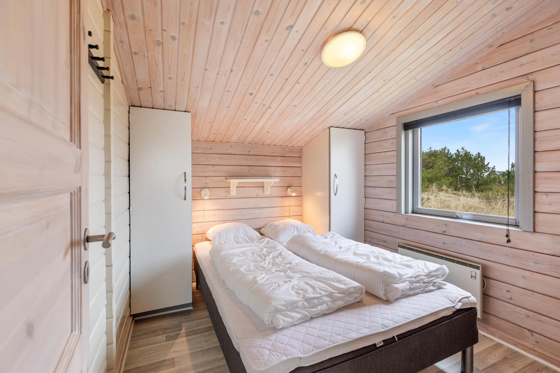 Un dormitorio con dos camas, paneles de madera clara y una ventana con vistas a la vegetación.