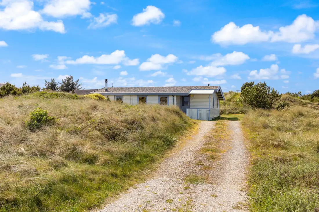 Una casa baja se encuentra al fondo de un camino arenoso entre dunas cubiertas de hierba.