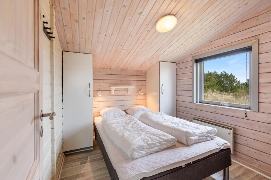 Un dormitorio luminoso con dos camas, paredes de madera clara y una ventana con vistas a los árboles.