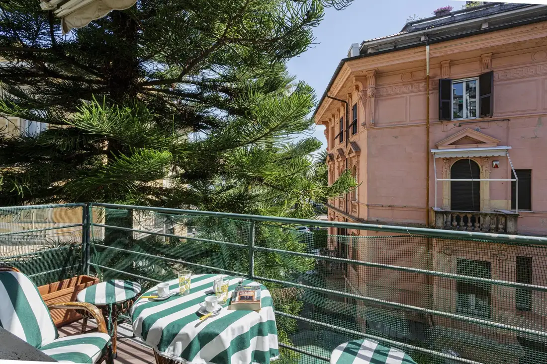 A balcony dining area with striped tablecloths overlooks trees and a pink building.